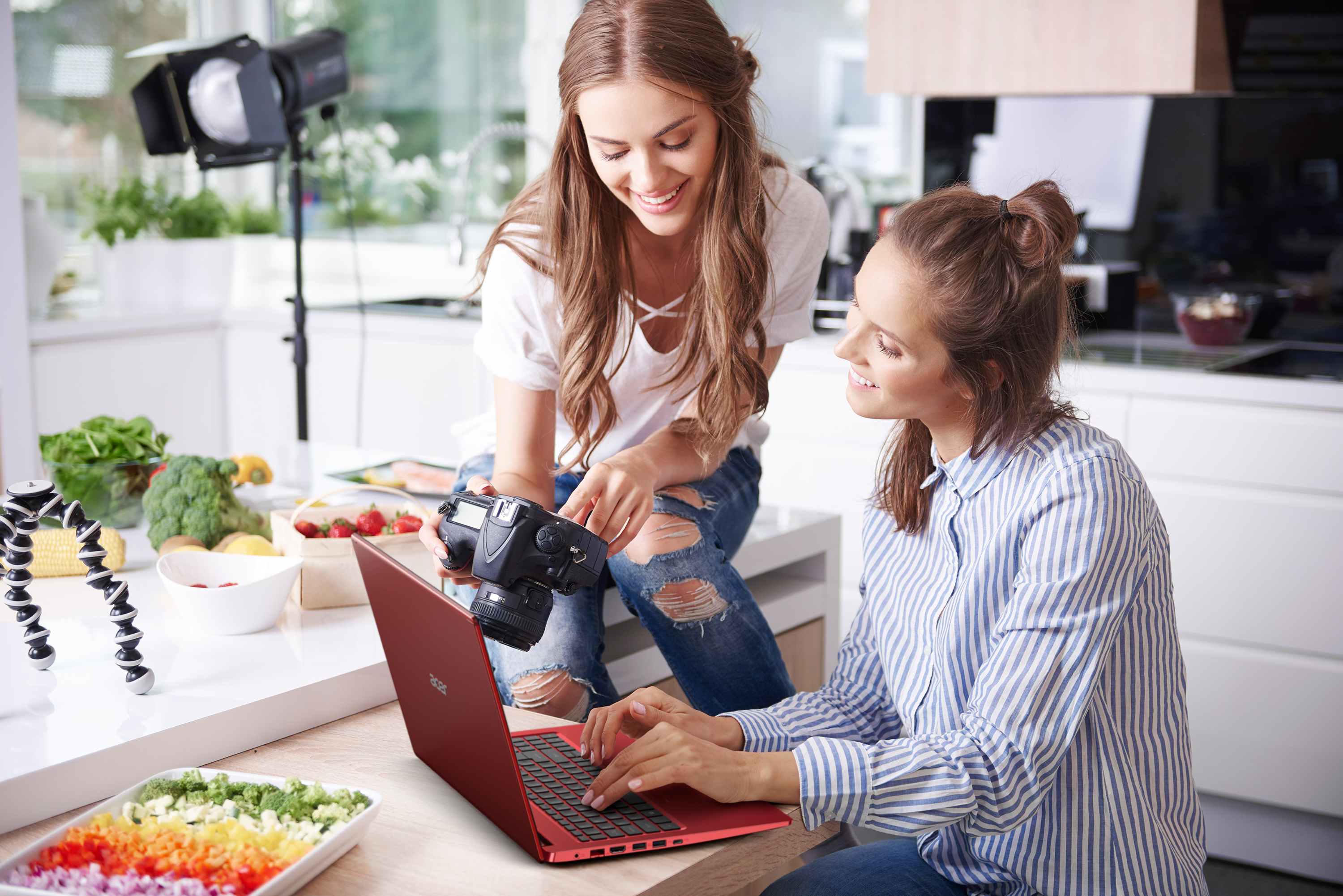 Two women -- one holding a camera -- working over a red Aspire 5 laptop in a kitchen with mise-en-place