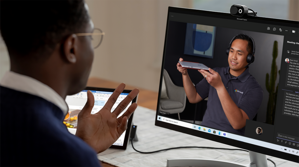 One man looks at a computer monitor where another man wearing a set of headphones is holding up a mobile device