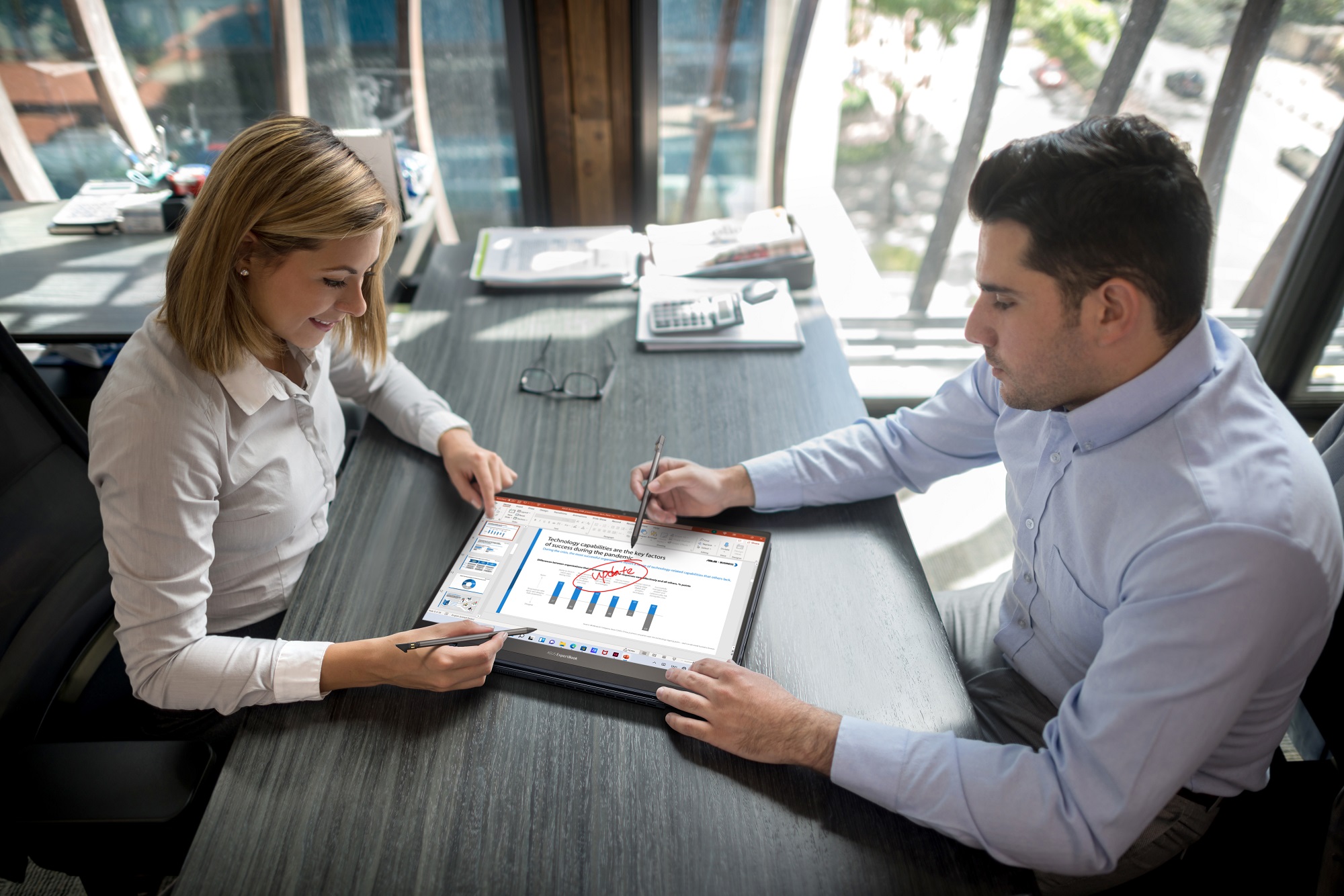 Two people sitting at a conference room table marking up a laptop screen with digital pens