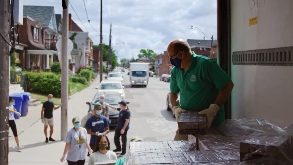 Man in mask passing out cans of food from back of a truck to people on the street in a neighborhood