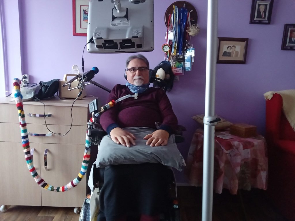 Alper Kaya sitting in a chair looking directly at camera, with a medical tube in his neck