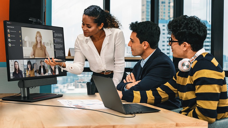 Three people in an office having a meeting with others shown on a display